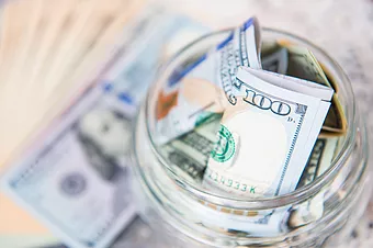 A variety of bills in a glass mason jar. A hundred dollar bill is the most prominent.